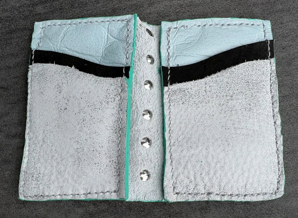 Wallet opened flat. The rivets from the back of the spikes are visible down the spine. Each side of the wallet has a bottom white/silver pocket, a higher black pocket, and light blue crocodile-embossed lining visible above that. Each side is stitched with silver thread.